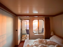 A cozy room with a neatly made bed and a window offering a view of a vibrant sunset over the ocean. Soft lighting creates a warm and inviting atmosphere, contrasting with the deep blue of the ocean. The room is simple, featuring neutral-colored walls and a curtain that partially covers the window, enhancing the intimate setting.