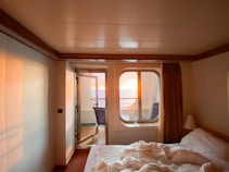 A cozy room with a neatly made bed and a window offering a view of a vibrant sunset over the ocean. Soft lighting creates a warm and inviting atmosphere, contrasting with the deep blue of the ocean. The room is simple, featuring neutral-colored walls and a curtain that partially covers the window, enhancing the intimate setting.
