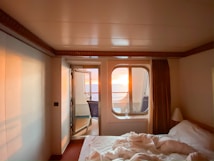 A cozy room with a neatly made bed and a window offering a view of a vibrant sunset over the ocean. Soft lighting creates a warm and inviting atmosphere, contrasting with the deep blue of the ocean. The room is simple, featuring neutral-colored walls and a curtain that partially covers the window, enhancing the intimate setting.