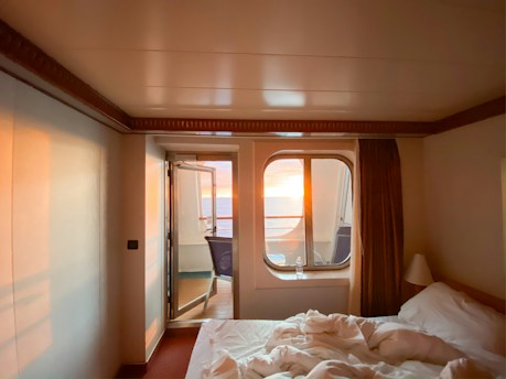 A cozy room with a neatly made bed and a window offering a view of a vibrant sunset over the ocean. Soft lighting creates a warm and inviting atmosphere, contrasting with the deep blue of the ocean. The room is simple, featuring neutral-colored walls and a curtain that partially covers the window, enhancing the intimate setting.