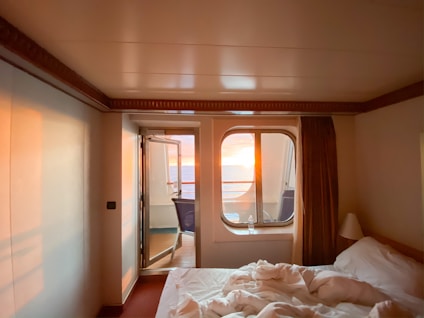 A cozy room with a neatly made bed and a window offering a view of a vibrant sunset over the ocean. Soft lighting creates a warm and inviting atmosphere, contrasting with the deep blue of the ocean. The room is simple, featuring neutral-colored walls and a curtain that partially covers the window, enhancing the intimate setting.