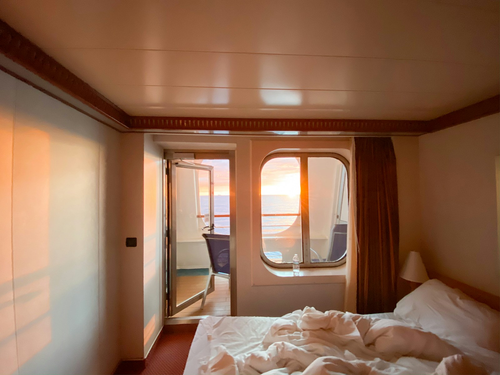 A ship's cabin with an unmade bed, a round porthole and an open balcony door, at orange sunset over the sea