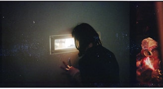 A person peers through a small rectangular window set in a dark wall. The light from the window illuminates the person's face partially, creating a sense of curiosity and intrigue. To the right, unfocused and colorful objects are visible, possibly reflecting items on the other side of the window.