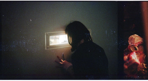 A person peers through a small rectangular window set in a dark wall. The light from the window illuminates the person's face partially, creating a sense of curiosity and intrigue. To the right, unfocused and colorful objects are visible, possibly reflecting items on the other side of the window.