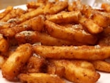 A close-up view of crispy, golden-brown seasoned fries, richly coated in spices and seasoning. The fries are piled together on a surface, showcasing a variety of textures and light reflections on their crispy edges.