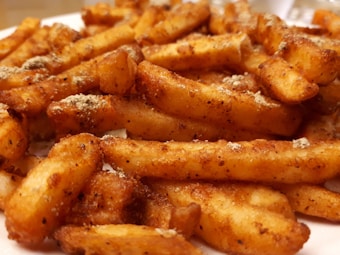 A close-up view of crispy, golden-brown seasoned fries, richly coated in spices and seasoning. The fries are piled together on a surface, showcasing a variety of textures and light reflections on their crispy edges.