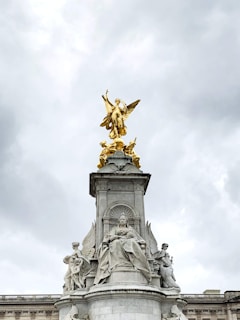 A grand monument featuring a prominent golden statue atop a pedestal surrounded by intricately carved stone figures. The central statue depicts a gilded angel-like figure raised high against a cloudy sky, with other detailed sculptures at lower levels, including a seated majestic figure.