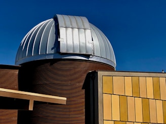 A modern observatory structure with a prominent metallic dome featuring a slit for telescopic observations. The building combines corrugated metal textures with a grid of light yellow and beige panels. The sky is clear and deep blue, offering a striking contrast to the structure.