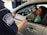 A border protection officer hands a document to a woman sitting in a car. Beside her is a black and white dog with its tongue out, looking friendly. The officer is in uniform, visible through the car window.