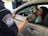 A border protection officer hands a document to a woman sitting in a car. Beside her is a black and white dog with its tongue out, looking friendly. The officer is in uniform, visible through the car window.