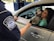A border protection officer hands a document to a woman sitting in a car. Beside her is a black and white dog with its tongue out, looking friendly. The officer is in uniform, visible through the car window.