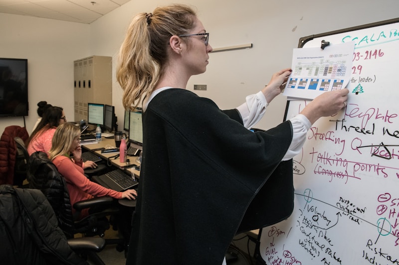 A woman presents information in an office while colleagues work at their desks.