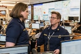 Two people in navy blue uniforms are engaged in conversation in a busy office environment, surrounded by multiple computer monitors displaying various maps and data. The setting appears to be a command or operations center with personnel focused on their screens.