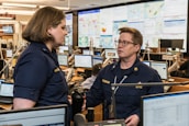 Two people in navy blue uniforms are engaged in conversation in a busy office environment, surrounded by multiple computer monitors displaying various maps and data. The setting appears to be a command or operations center with personnel focused on their screens.