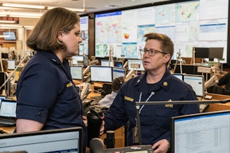 Two people in navy blue uniforms are engaged in conversation in a busy office environment, surrounded by multiple computer monitors displaying various maps and data. The setting appears to be a command or operations center with personnel focused on their screens.