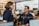 Two people in navy blue uniforms are engaged in conversation in a busy office environment, surrounded by multiple computer monitors displaying various maps and data. The setting appears to be a command or operations center with personnel focused on their screens.