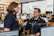 Two people in navy blue uniforms are engaged in conversation in a busy office environment, surrounded by multiple computer monitors displaying various maps and data. The setting appears to be a command or operations center with personnel focused on their screens.
