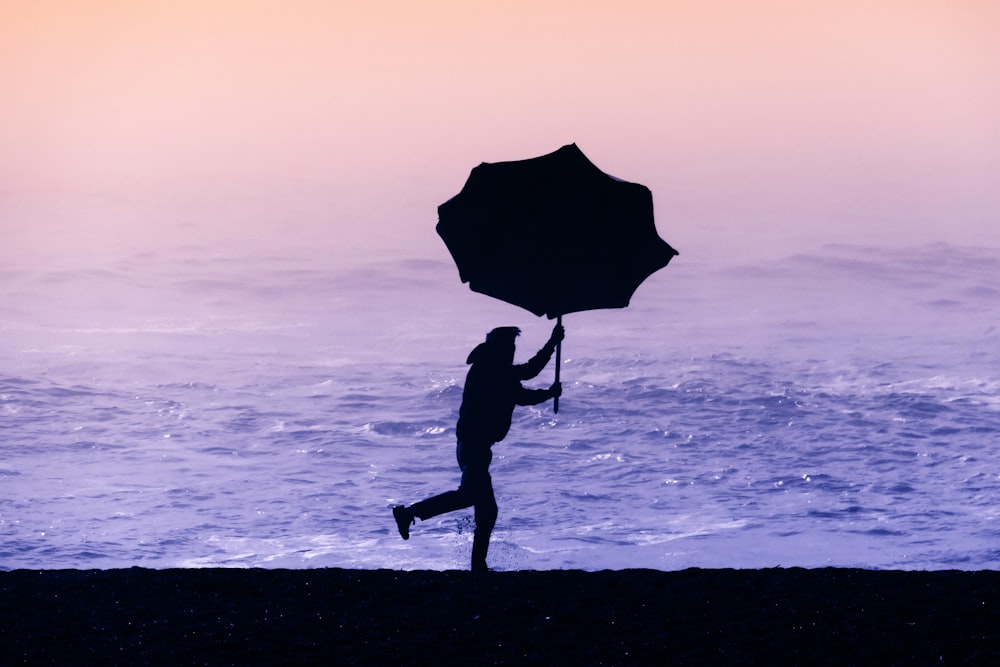 silhouette of person holding umbrella walking on seashore during sunset