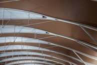 A modern architectural ceiling design with a patterned arrangement of wood and white translucent panels. The structure features curved beams and sleek metal supports, creating a sense of spaciousness and symmetry.