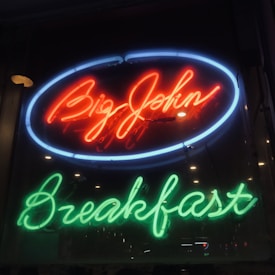 A neon sign featuring the words 'Big John Breakfast'. The sign is lit in vibrant colors, with 'Big John' in bright red housed within a blue oval, and 'Breakfast' in green below it. There are reflections of city lights visible in the background.