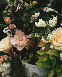 A bouquet of various flowers including roses, carnations, and small white blossoms, surrounded by lush green foliage. The arrangement is dense and colorful, with a mix of textures and sizes.
