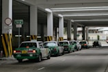 A row of green taxis are lined up under a covered structure, waiting in a designated taxi stand area. The environment has industrial features with pillars and signage in both English and another language. The concrete surface and overhead fluorescent lighting contribute to a utilitarian atmosphere.
