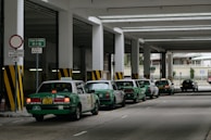 A row of green taxis are lined up under a covered structure, waiting in a designated taxi stand area. The environment has industrial features with pillars and signage in both English and another language. The concrete surface and overhead fluorescent lighting contribute to a utilitarian atmosphere.