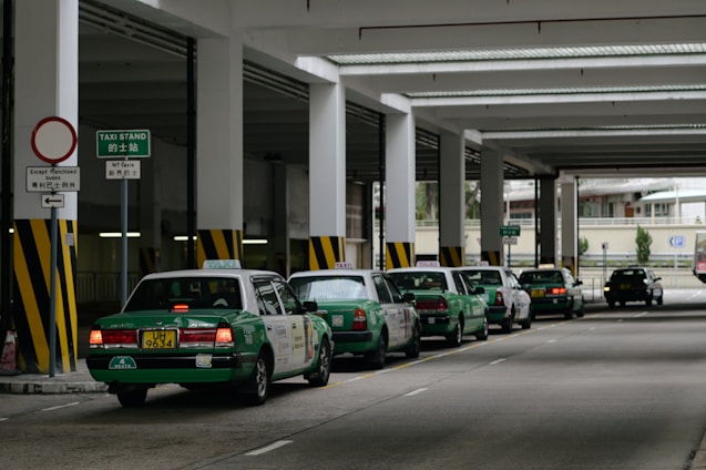 A row of green taxis are lined up under a covered structure, waiting in a designated taxi stand area. The environment has industrial features with pillars and signage in both English and another language. The concrete surface and overhead fluorescent lighting contribute to a utilitarian atmosphere.