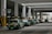 A row of green taxis are lined up under a covered structure, waiting in a designated taxi stand area. The environment has industrial features with pillars and signage in both English and another language. The concrete surface and overhead fluorescent lighting contribute to a utilitarian atmosphere.