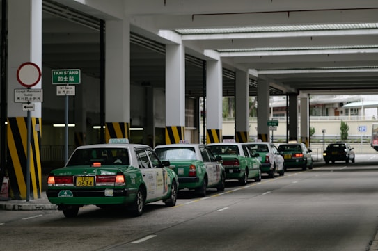 A row of green taxis are lined up under a covered structure, waiting in a designated taxi stand area. The environment has industrial features with pillars and signage in both English and another language. The concrete surface and overhead fluorescent lighting contribute to a utilitarian atmosphere.