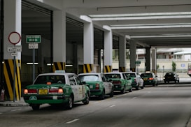 A row of green taxis are lined up under a covered structure, waiting in a designated taxi stand area. The environment has industrial features with pillars and signage in both English and another language. The concrete surface and overhead fluorescent lighting contribute to a utilitarian atmosphere.
