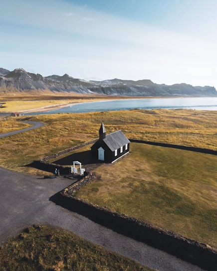 A small black church with a white door and windows is situated in a rural landscape. It is surrounded by a stone wall and set against a backdrop of gently rolling hills and distant snow-capped mountains. A winding road leads to the church, and the weather is clear with bright sunlight casting shadows on the ground.