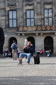 A street musician is playing a clarinet while seated on a folding chair near a historic building. People are sitting and standing nearby, some admiring the performance. The building's facade features large windows and ornate ironwork.