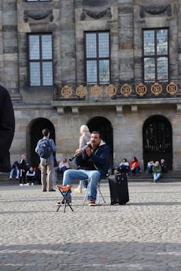 A street musician is playing a clarinet while seated on a folding chair near a historic building. People are sitting and standing nearby, some admiring the performance. The building's facade features large windows and ornate ironwork.