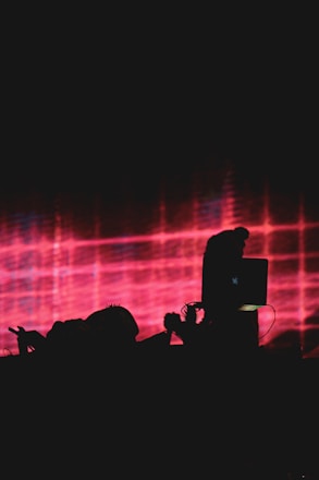 A dark silhouette of a person working on a laptop is set against a vibrant backdrop of red and pink grid patterns. The scene suggests a concert or performance setting.