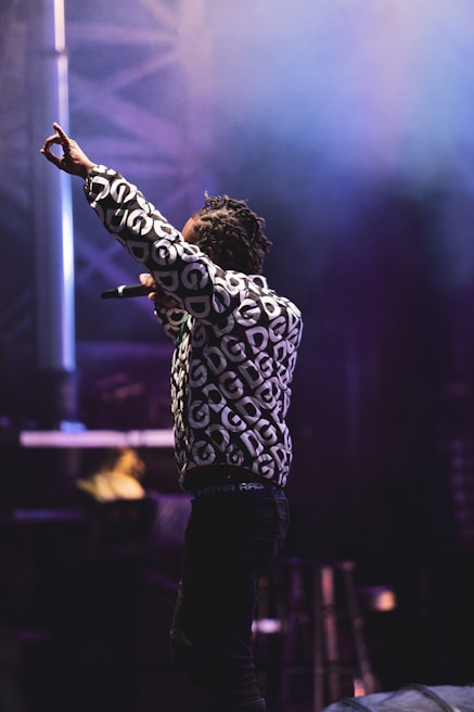 A person is performing on stage, wearing a patterned jacket with a design of repeating letters. The background is filled with stage lights and a misty atmosphere, creating a concert-like setting. The person is holding a microphone and raising one arm into the air.