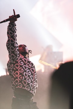A performer wearing a patterned jacket with initials raises their hand holding a microphone on stage, surrounded by bright lights and a smoke-filled ambiance. A blurred background suggests a DJ setup.