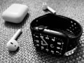 A black smartwatch with a perforated strap is placed on a textured surface alongside wireless earbuds and their charging case. The watch displays an analog clock face, and the earbuds are positioned with one lying horizontally and the other propped against the case.
