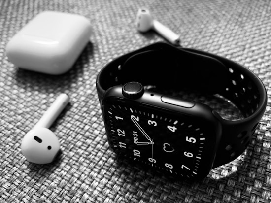 A black smartwatch with a perforated strap is placed on a textured surface alongside wireless earbuds and their charging case. The watch displays an analog clock face, and the earbuds are positioned with one lying horizontally and the other propped against the case.