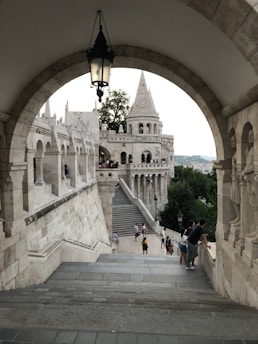 An architectural scene featuring an arched stone structure leading to a castle-like building with multiple spires. The foreground includes stone steps and a hanging lantern, while the background showcases greenery and a view of more stone arches. Several people are casually walking and standing around, some gazing over the edge, suggesting tourism or leisurely sightseeing.