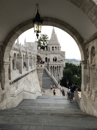 An architectural scene featuring an arched stone structure leading to a castle-like building with multiple spires. The foreground includes stone steps and a hanging lantern, while the background showcases greenery and a view of more stone arches. Several people are casually walking and standing around, some gazing over the edge, suggesting tourism or leisurely sightseeing.
