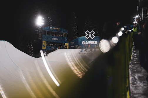 A nighttime scene at the X Games featuring a snowboarding half-pipe under bright floodlights. The ESPN booth is visible in the background along with banners and some spectators. A long, blurred line of lights adds a dynamic effect along the edge of the slope.