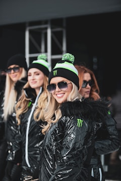 A group of smiling women standing together, wearing shiny black jackets and black beanies with green accents. They are also wearing sunglasses and appear to be posing for the camera. The background includes a metal structure, suggesting an outdoor or event setting.