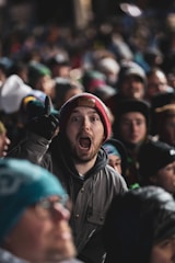 A large crowd of people is gathered, with one person in the forefront wearing a red beanie and expressing excitement or enthusiasm. The person's mouth is open and they appear to be shouting or cheering, surrounded by others wearing winter clothing, suggesting a chilly environment.