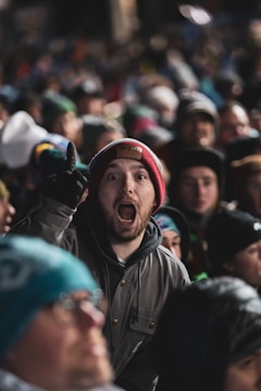 A large crowd of people is gathered, with one person in the forefront wearing a red beanie and expressing excitement or enthusiasm. The person's mouth is open and they appear to be shouting or cheering, surrounded by others wearing winter clothing, suggesting a chilly environment.