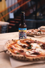 A bottle of pilsner beer with a colorful, artistic label is placed on a wooden table next to a freshly baked pizza. The pizza has a golden crust, melted cheese, and visible toppings such as basil and tomato sauce. The background suggests an indoor setting with blurred stairs and a cozy ambiance.