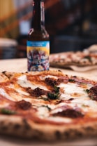 A close-up view of a freshly baked pizza with melted cheese, fresh basil leaves, and dollops of tomato sauce topping. In the background, there's a bottle of craft beer with a colorful label showing a lively design. The background appears to be out of focus, highlighting the pizza and beer bottle.