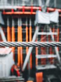 A close-up of a thick metal wire is in focus with a blurred construction site in the background. The construction site features orange and gray scaffolding and materials.