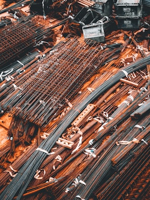 A collection of metal rods and bars are organized on the ground, tied together with what appear to be white straps. Some of these metal pieces form grid-like structures, likely reinforcing wire meshes. The setting conveys a scene typical of a construction site, where materials are being prepared or stored for use.