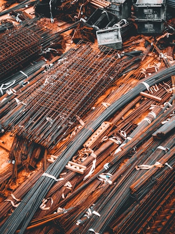 A collection of metal rods and bars are organized on the ground, tied together with what appear to be white straps. Some of these metal pieces form grid-like structures, likely reinforcing wire meshes. The setting conveys a scene typical of a construction site, where materials are being prepared or stored for use.